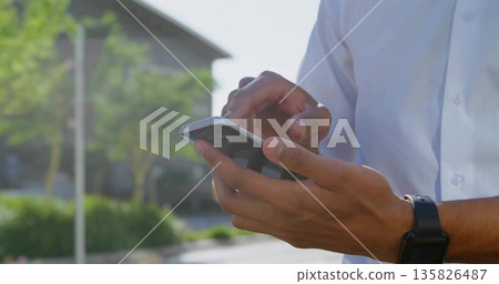 Tapping smartphone, man in white shirt and black smartwatch standing near parking, blurred trees 135826487