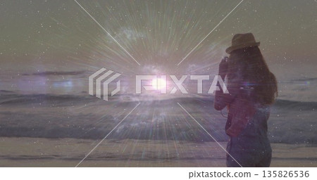 Pointing camera, woman wearing fedora capturing cosmic light over ocean waves at shore, copy space Pointing camera, woman wearing fedora capturing cosmic light over ocean waves at shore, copy space 135826536
