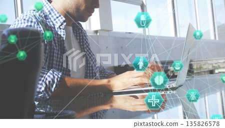 Typing Hispanic man in checkered shirt at glass desk, with silver laptop and floating digital icons 135826578