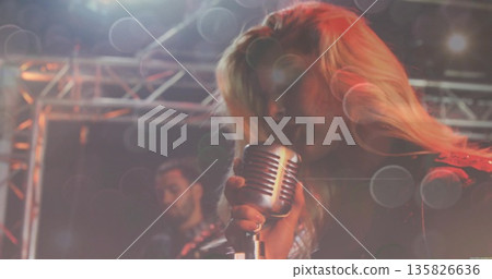 Singing female wearing stage attire holding retro microphone on stage with guitar and lighting rig Singing female wearing stage attire holding retro microphone on stage with guitar and lighting rig 135826636