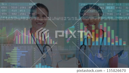 Standing two doctors wearing lab coat and scrubs examining data charts in clinic, with stethoscopes Standing two doctors wearing lab coat and scrubs examining data charts in clinic, with stethoscopes 135826670