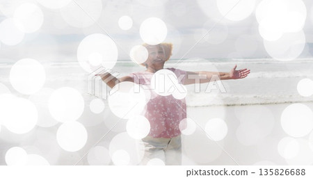 Senior woman in floral blouse stretching arms wide on shore by ocean waves, with bokeh overlay Senior woman in floral blouse stretching arms wide on shore by ocean waves, with bokeh overlay 135826688