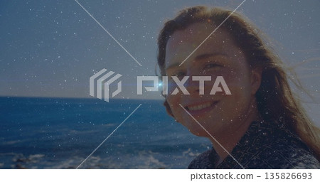 Standing woman in geometric shirt smiling at camera on beach edge, sunlight reflecting off water 135826693