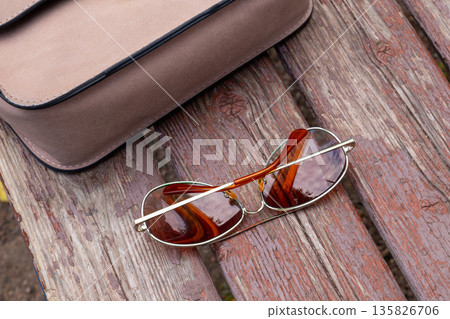 Aviator sunglasses with large drop shaped lenses and thin metal frame. Sunglasses and handbag on rustic wooden bench. Leisure and relaxation. Aviator sunglasses with large drop shaped lenses and thin metal frame. Sunglasses and handbag on rustic wooden bench. Leisure and relaxation. 135826706