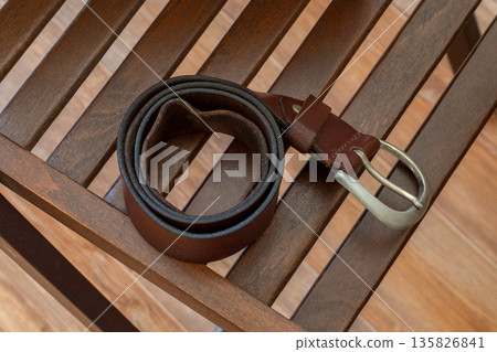 Brown leather belt with a silver buckle on a wooden slatted chair, showing fashion accessory and casual style. Brown leather belt with a silver buckle on a wooden slatted chair, showing fashion accessory and casual style. 135826841