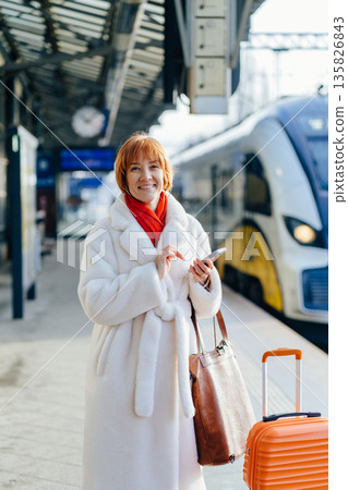 Smiling woman in white coat holding smartphone and leather bag, standing on railway platform with orange suitcase. Rail station, positive travel, business trip, public transport, confident passenger. Smiling woman in white coat holding smartphone and leather bag, standing on railway platform with orange suitcase. Rail station, positive travel, business trip, public transport, confident passenger. 135826843
