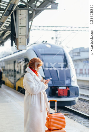 Middle aged Caucasian woman checking smartphone while standing near train, stacked orange suitcases on railway platform. Concept of ecology, work travel, business, everyday rail commuting in Europe. Middle aged Caucasian woman checking smartphone while standing near train, stacked orange suitcases on railway platform. Concept of ecology, work travel, business, everyday rail commuting in Europe. 135826853