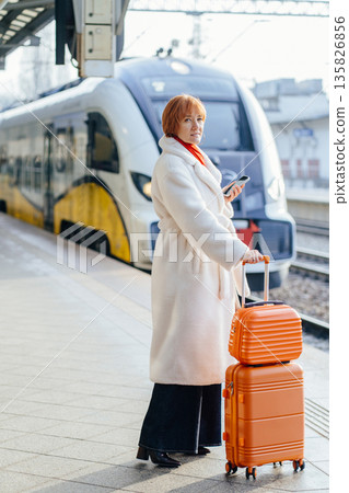 Caucasian woman in white winter coat with red scarf standing on railway platform, holding smartphone, waiting for regional train. European rail travel, business trip, public transport routine. Caucasian woman in white winter coat with red scarf standing on railway platform, holding smartphone, waiting for regional train. European rail travel, business trip, public transport routine. 135826856
