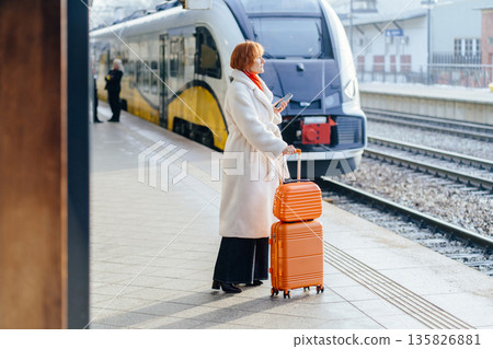 Mature Caucasian woman standing on railway platform beside orange suitcase, holding smartphone and looking toward arriving electric train. Professional routine, public transit, winter city journey. Mature Caucasian woman standing on railway platform beside orange suitcase, holding smartphone and looking toward arriving electric train. Professional routine, public transit, winter city journey. 135826881