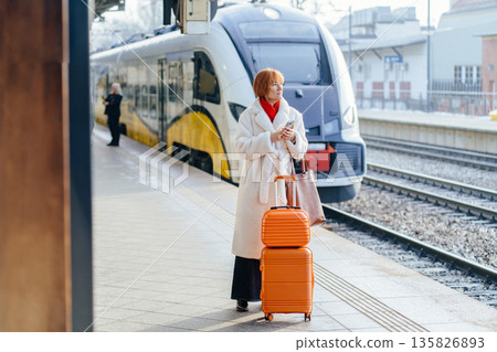 Mature Caucasian woman standing on railway platform with orange suitcase and leather bag, checking smartphone near modern electric train. Work travel, business trip, rail transport concept 135826893