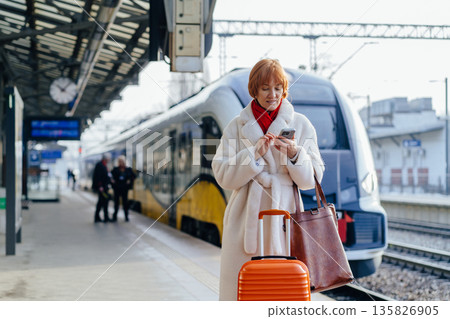 Smiling European woman traveler with suitcase standing at railway station platform, lady ready for weekend trip train stands on platform in background, beginning of journey Smiling European woman traveler with suitcase standing at railway station platform, lady ready for weekend trip train stands on platform in background, beginning of journey 135826905