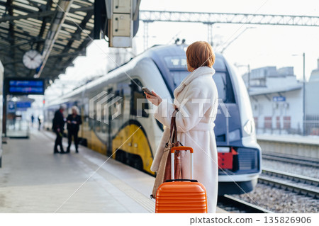 Rear view of elegant woman with a suitcase and a backpack standing at the train station waiting for train, using a mobile phone. Business trip, work travel, public transport, solo female passenger Rear view of elegant woman with a suitcase and a backpack standing at the train station waiting for train, using a mobile phone. Business trip, work travel, public transport, solo female passenger 135826906