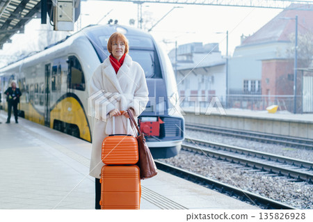 Serious caucasian business traveler standing on train platform with orange suitcase. Rail mobility, professional relocation, intercity commute, modern public transport in everyday city life. Serious caucasian business traveler standing on train platform with orange suitcase. Rail mobility, professional relocation, intercity commute, modern public transport in everyday city life. 135826928