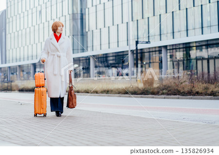Independent adult woman walks alone with suitcase along modern city street. Urban mobility, relocation journey, solo lifestyle, confident movement, everyday travel supported by digital tools. 135826934