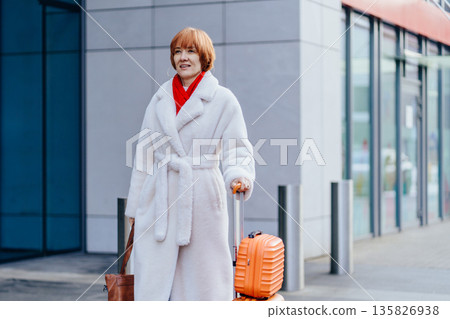 Confident red-haired mature Caucasian woman walking outdoors with suitcase and smartphone near modern building. Relocation , solo travel, lifestyle, everyday movement, digital navigation, city commute Confident red-haired mature Caucasian woman walking outdoors with suitcase and smartphone near modern building. Relocation , solo travel, lifestyle, everyday movement, digital navigation, city commute 135826938