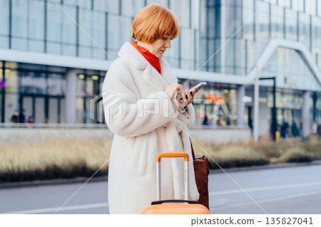 Mature woman using smartphone while standing with suitcase on city street near public building. Concept of checking, coordination relocation, urban mobility, travel management, digital navigation. 135827041