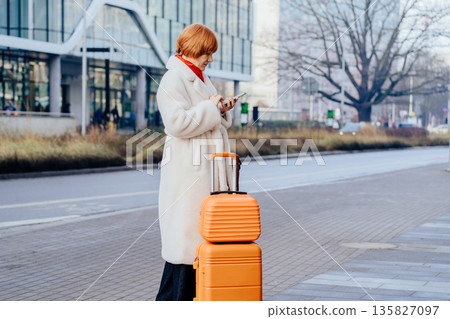 Mature woman using smartphone while standing with luggage on city. Concept of digital coordination, checking directions, urban mobility, relocation, everyday travel management, transit routine. Mature woman using smartphone while standing with luggage on city. Concept of digital coordination, checking directions, urban mobility, relocation, everyday travel management, transit routine. 135827097