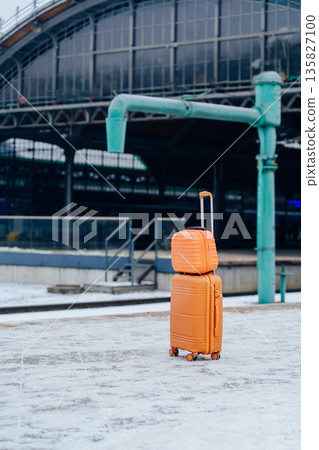Vertical of orange suitcase standing alone on railway station platform in winter. Concept of travel, relocation, departure pause, transit moment, mobility, public transport infrastructure. 135827100