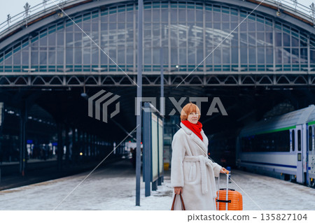 Mature woman waiting with suitcase on railway station platform. Concept of urban transit space, relocation moment, winter travel routine, mobility, public infrastructure, personal journey. Mature woman waiting with suitcase on railway station platform. Concept of urban transit space, relocation moment, winter travel routine, mobility, public infrastructure, personal journey. 135827104