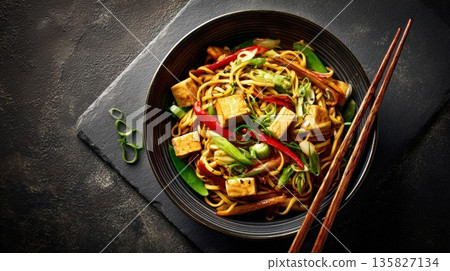 Tofu noodle stir-fry is served in a dark bowl with chopsticks 135827134