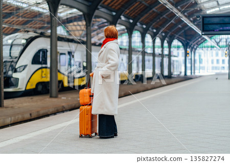 Full height of redhead Caucasian middle age woman in white coat standing on railway platform with suitcase, waiting electric train, calm urban mobility moment, sustainable public transport choice. Full height of redhead Caucasian middle age woman in white coat standing on railway platform with suitcase, waiting electric train, calm urban mobility moment, sustainable public transport choice. 135827274