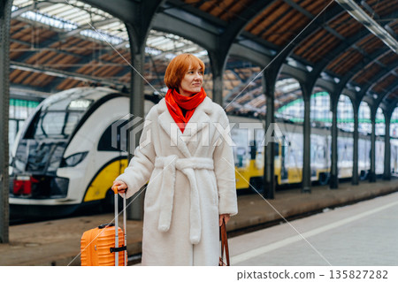 Redhead middle age woman in white coat pulling suitcase on railway station platform, choosing electric train, daily mobility, eco friendly transport, urban relocation, sustainable city in Europe Redhead middle age woman in white coat pulling suitcase on railway station platform, choosing electric train, daily mobility, eco friendly transport, urban relocation, sustainable city in Europe 135827282