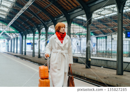 Thoughtful mature woman in white faux fur coat and red scarf standing with orange suitcase on railway platform of railway station. Winter fashion, modern city lifestyle, contemporary travel aesthetic. Thoughtful mature woman in white faux fur coat and red scarf standing with orange suitcase on railway platform of railway station. Winter fashion, modern city lifestyle, contemporary travel aesthetic. 135827285