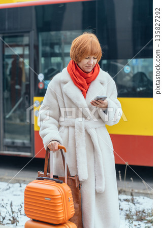 Vertical of red haired woman with luggage using smartphone in snowy European city. Holiday vacation planning, urban travel, solo female traveler checking phone during winter trip abroad. Vertical of red haired woman with luggage using smartphone in snowy European city. Holiday vacation planning, urban travel, solo female traveler checking phone during winter trip abroad. 135827292
