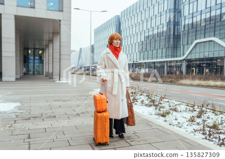 Full body mature woman walking outdoors with orange suitcase near modern glass office buildings. Urban travel, relocation, business trip, solo female traveler, winter, architecture, mobility concept. Full body mature woman walking outdoors with orange suitcase near modern glass office buildings. Urban travel, relocation, business trip, solo female traveler, winter, architecture, mobility concept. 135827309