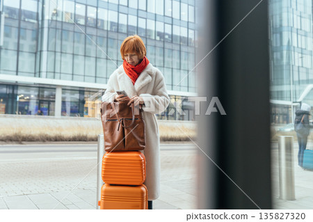 Mature woman holding smartphone and standing with suitcase near office building. Concept of business travel, transport delay, arrival, independent female traveler in city environment. 135827320