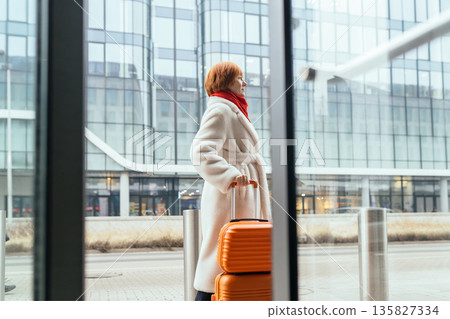 Mature woman in light coat standing outdoors with orange suitcase near modern glass building. Concept of travel, relocation, business trip, urban mobility, independence, solo traveler in city 135827334