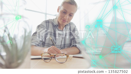 Writing woman in plaid top sitting at desk in office with notebook succulent laptop network overlay Writing woman in plaid top sitting at desk in office with notebook succulent laptop network overlay 135827502
