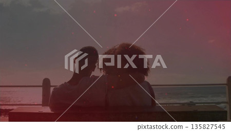 Sitting couple watching ocean at sunset on wooden bench by metal railing, featuring bag with straps 135827545
