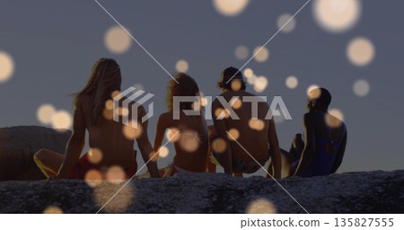 Watching friends wearing swimwear sitting on rocky ledge at dusk, with bokeh light orbs 135827555