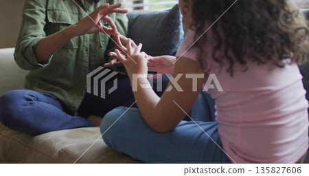 Communicating mother and daughter sitting legs crossed on sofa at home with pillow and blinds 135827606