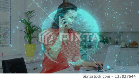 Talking woman in red blouse using smartphone typing on laptop in kitchen, with data sphere overlay 135827644