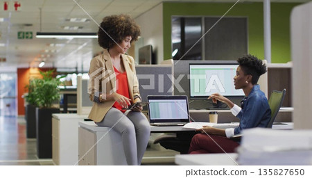Discussing two colleagues in business casual sharing data on tablet and laptops in open-plan office Discussing two colleagues in business casual sharing data on tablet and laptops in open-plan office 135827650