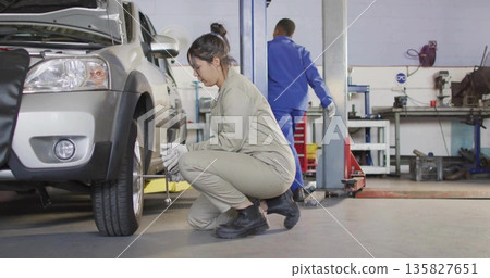 Crouching female mechanic removing lug nuts on shop floor beneath hydraulic lift, with lug wrench Crouching female mechanic removing lug nuts on shop floor beneath hydraulic lift, with lug wrench 135827651