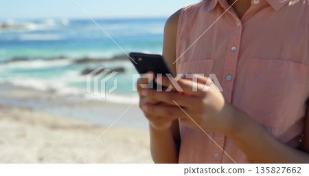 Standing woman wearing pink sleeveless shirt at beach shoreline, holding black smartphone Standing woman wearing pink sleeveless shirt at beach shoreline, holding black smartphone 135827662