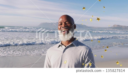 Smiling senior man standing on sandy shoreline by ocean waves, with floating golden star overlays Smiling senior man standing on sandy shoreline by ocean waves, with floating golden star overlays 135827814