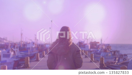 Gazing woman wearing hoodie, trousers facing fishing boats on harbor pier, with bollards and ropes 135827833