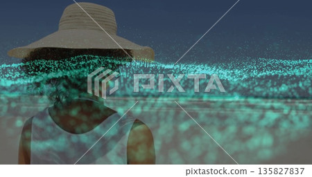 Standing woman wearing straw hat, white top at dusk beach, with glowing turquoise particle overlay Standing woman wearing straw hat, white top at dusk beach, with glowing turquoise particle overlay 135827837