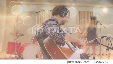 Male guitarist wearing plaid shirt, playing acoustic guitar in recording studio, with red drum kit 135827870