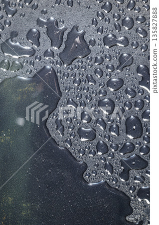 An abstract macro image showing clear water droplets and puddles of various sizes on a dark, grainy, charcoal-colored surface with light reflections An abstract macro image showing clear water droplets and puddles of various sizes on a dark, grainy, charcoal-colored surface with light reflections 135827888