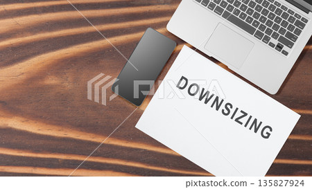 Top-down view of a modern workspace featuring a laptop, smartphone, and a paper with the text DOWNSIZING representing corporate layoffs and restructuring Top-down view of a modern workspace featuring a laptop, smartphone, and a paper with the text DOWNSIZING representing corporate layoffs and restructuring 135827924