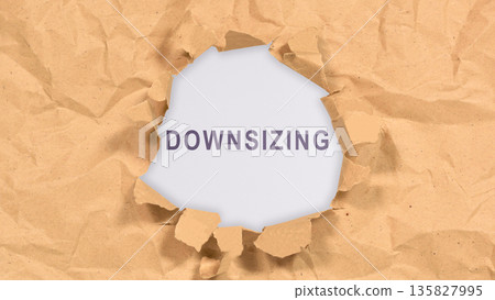 An image showing the word 'DOWNSIZING' visible through a ragged hole in a sheet of crumpled brown kraft paper, symbolizing business restructuring and layoffs 135827995