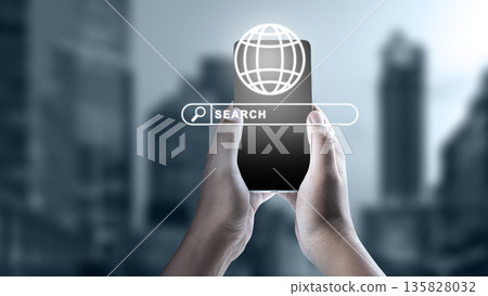 Hands holding a mobile phone with a digital search bar and globe icon against a blurred urban background. Concept of global information technology, connectivity, and internet search 135828032