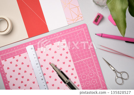 Pink sheet with ruler and knife on cutting mat among craft tools and colored paper. Creative workspace for art and design preparation. Pink sheet with ruler and knife on cutting mat among craft tools and colored paper. Creative workspace for art and design preparation. 135828527
