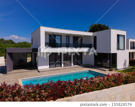 Modern luxury villa with swimming pool and clean minimalist architecture under a blue sky. Contemporary residential exterior featuring large windows, terrace, and elegant outdoor living space Modern luxury villa with swimming pool and clean minimalist architecture under a blue sky. Contemporary residential exterior featuring large windows, terrace, and elegant outdoor living space 135828574