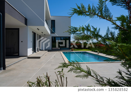 Modern luxury villa with swimming pool and clean minimalist architecture under a blue sky. Contemporary residential exterior featuring large windows, terrace, and elegant outdoor living space 135828582
